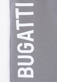 Pantaloni della tuta grigi con audace testo bianco "BUGATTI" sulla gamba sinistra. Il tessuto è liscio con una trama leggermente elasticizzata e un design aderente.