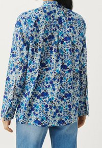 Woman wearing blue floral pattern long-sleeve blouse and light blue jeans, shown from the back against a plain background.
