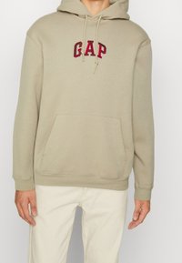 Sudadera con capucha de algodón beige con bolsillo tipo canguro en el frente y logo "GAP" bordado en rojo. Incluye capucha con cordón ajustable y puños acanalados.