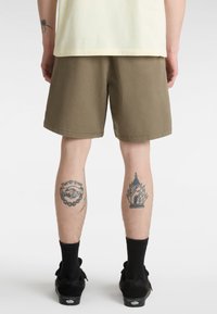 Khaki-Shorts im entspannten Schnitt aus Baumwollstoff mit aufgerolltem Saum. Sichtbare schwarze Socken und Schuhe sowie Tattoos an den Beinen.