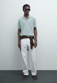 Mintgrön polo-skorta, vita raka byxor, brun tröja knuten i midjan, svarta solglasögon och mörka skor på en strukturerad yta.