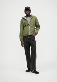 Veste légère olive avec capuche, fermeture éclair et poignets élastiques, assortie à un jean noir à grandes jambes et des baskets noires.