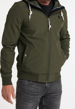 Homme portant une veste zippée vert olive avec des cordons, des poches à boutons et des poignets côtelés, associée à un jean gris foncé.
