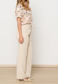 Top leggero a maniche corte di colore rosa chiaro con motivo di paillettes floreali, abbinato a pantaloni beige a vita alta e larghi, insieme a tacchi coordinati.