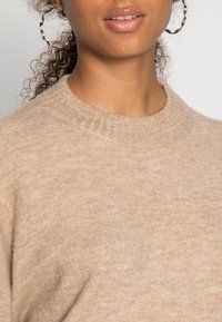 Beige Strickpullover mit einem gerippten Rundhalsausschnitt, weicher Textur und subtilen Farbvariationen. Hat einen figurbetonten Schnitt.