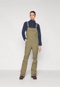 Olivgröna byxor med justerbara remmar, en framficka och dragkedjor på sidorna; kombinerat med en marinblå fleece-tröja och svarta skor.