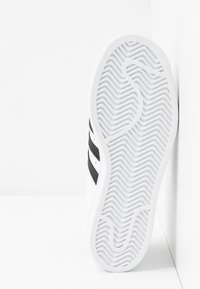 Baskets blanches avec un design à trois bandes noires. Elles possèdent une semelle en caoutchouc texturée avec un motif ondulé et une tige en matériau lisse.