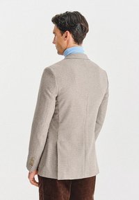 Beżowa marynarka w zygzaki z teksturowanej tkaniny, z klapami w kształcie notcha i zapięciami na guziki. Noszona na jasnoniebieskim golfie. Dopasowany krój.