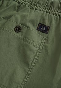 Pantalones cortos de tela verde con cintura elástica, que cuentan con un bolsillo lateral con un botón oscuro y una etiqueta azul con un logo blanco "M".
