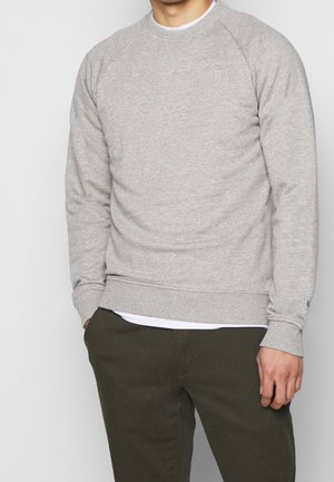 Uomo che indossa una felpa a maniche lunghe grigio chiaro sopra una camicia bianca, abbinata a pantaloni verde scuro, con le mani parzialmente in tasca.