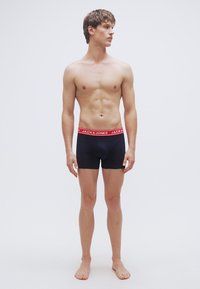 Μαύρα βαμβακερά μποξεράκια με κόκκινη ελαστική ζώνη μέσης που φέρει το λογότυπο "JACK & JONES", φορεμένα από έναν άνδρα μοντέλο που στέκεται μπροστά σε απλό φόντο.