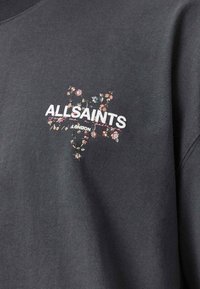 Σκούρο γκρι φούτερ με το κείμενο «ALLSAINTS LONDON» πάνω από ένα λουλουδένιο σχέδιο σε σχήμα σταυρού σε παστέλ χρώματα στο στήθος.