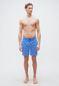 Νέος άνδρας με σγουρά μαλλιά που φοράει μπλε μαγιό με σχέδια λευκού αλόγου, στέκεται ξυπόλητος μπροστά σε ένα απλό λευκό φόντο.