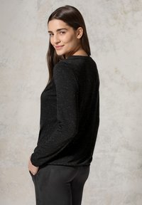 Schwarzes Langarmshirt mit dezenten Glitzerpartikeln, Rundhalsausschnitt und taillierter Passform. Das Model trägt dazu eine dunkle, weiche Stoffhose.