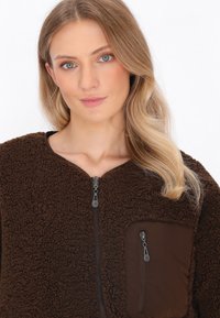 Giacca in pile marrone con una superficie testurizzata, chiusura frontale con zip e una tasca con zip marrone a contrasto sul petto sinistro.