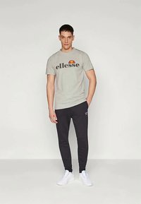 Pelēka kokvilnas T-kreklu ar melnu "ellesse" logo un sarkanu saules dizainu, kopā ar melnajām sporta biksēm un baltām čībām. Priekšējais skats, stāvoša pozīcija.