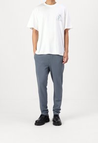 White cotton t-shirt with blue graphic on left chest, paired with gray jogger pants and black lace-up shoes. Simple, relaxed fit.
