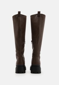 Bottes montantes marron en matériau synthétique lisse. Elles présentent une semelle noire épaisse, des détails zippés et des languettes de maintien en haut.