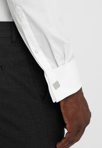 Weißes Hemd mit einem quadratischen silbernen Manschettenknopf, ordentlich gefaltet. Der Stoff des Hemdes hat eine glatte Textur und wird mit dunklen strukturierten Hosen kombiniert.