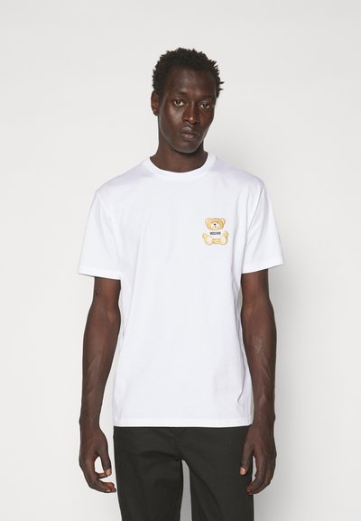 Camiseta de algodón blanca con un logo de osito de peluche amarillo bordado en el lado izquierdo del pecho, con mangas cortas y diseño de cuello redondo.