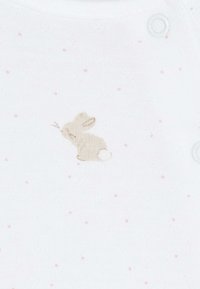 Combinaison en tissu blanc avec de petits pois beiges et une broderie de lapin. Elle présente une texture douce et une fermeture à pressions à l'épaule.