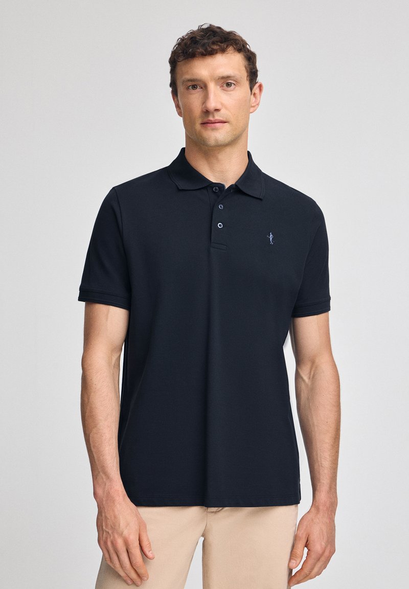 Mörkblå polo-skorta i slät väv med krage och tre-knapps knäppning; har en liten logotyp på vänster bröst.