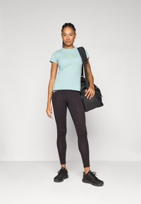 Lyseblå trenings T-skjorte med grønt logo, sorte leggings og svarte joggesko. Modellen bærer en svart treningsbag. Enkelt bakgrunn.