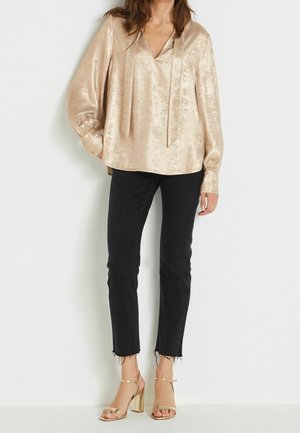 Femme portant un chemisier beige brillant à manches longues, un jean noir slim aux ourlets effilochés et des sandales dorées à talons hauts sur fond blanc.