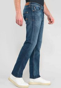 Jeans de denim en un lavado azul medio con corte recto. Las características incluyen cinco bolsillos, detalles de costura y un parche de cuero marrón en la cintura.
