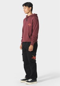 Felpa maroon con cappuccio, tasca frontale e dettagli grafici. Abbinata a pantaloni cargo neri e sneakers.
