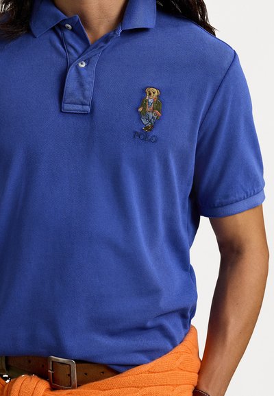 Zils polo krekls ar mīkstu tekstūru, ar izšūtu lāču dizainu un "Polo" tekstu uz krūtīm. Oranža džemperis sasiet ap vidukli.