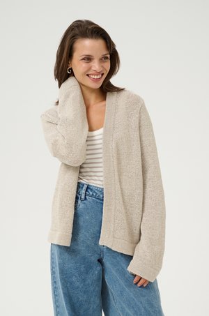 Cardigan en maille beige clair avec une ouverture sur le devant, des poignets côtelés et un tissu texturé, associé à un haut rayé et un jean en denim taille haute.