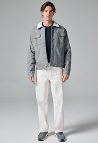 Grå denimjakke med en hvid shearlingkrave, frontlukning med knapper og to brystlommer, parret med hvide jeans og grå sneakers.