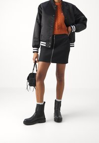 Sort bomberjakke med hvide stribede manchetter, orange kabelstrikket sweater, sort lynlås-skjørt, sorte chunky ankelstøvler og lille sort håndtaske.