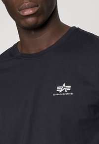 Marineblaue Baumwoll-T-Shirt mit Rundhalsausschnitt; verfügt über ein weißes Logo "ALPHA INDUSTRIES" mit grafischen Akzenten im linken Brustbereich.