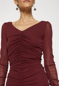 Burgundy dlouhé šaty s dlouhým rukávem, naškrobenou texturou a výstřihem do V. Průsvitné akcenty na rukávech zvýrazňují design.