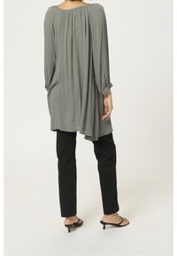 Túnica de manga larga, de corte holgado, en suave tejido gris apagado, con un respaldo con pliegues, combinada con pantalones negros de corte ajustado y sandalias negras.