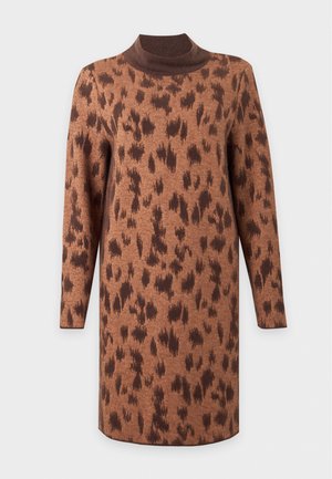 Une femme porte une robe en maille marron à motifs, un col roulé foncé et des bottines noires, sur un fond clair uni.