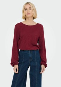 Top lungo a maniche lunghe in borgogna con scollo rotondo, abbinato a jeans a gamba larga di colore blu scuro a vita alta con cuciture verticali.