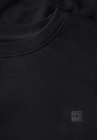 Svart sweatshirt med rund ribbad halsringning och en texturerad logotagg på framsidan. Tyget verkar slätt och enhetligt i textur.