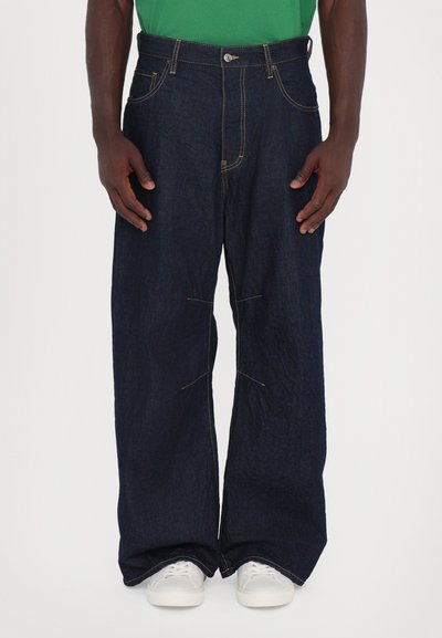 Jean large en denim foncé avec des coutures jaunes contrastantes, cinq poches et une fermeture à bouton à l'avant, associés à des baskets blanches.