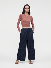 Femme debout sur un fond uni, portant un cardigan court boutonné rose poudré, un pantalon large bleu foncé à motifs, et des sandales marron.