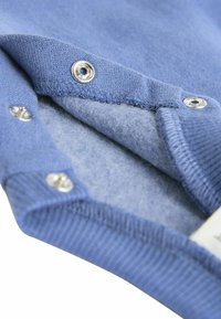 Zils džemperis ar mīkstu iekšpusi, uzkurināts ar sudraba pogām, ribotu piedurkņu dizainu un teksturētu audumu. Redzētā detaļa rāda iekšējo oderi.