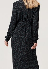 Femme aux cheveux blonds portant une longue robe noire à imprimé floral bleu, manches longues et ceinture à la taille, vue de dos.