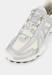 Białe siatkowe trampki z srebrnymi akcentami, charakteryzujące się teksturowanym wzorem, okrągłymi sznurówkami i wyściełaną podeszwą. Kątowe wycięcia poprawiają wentylację.