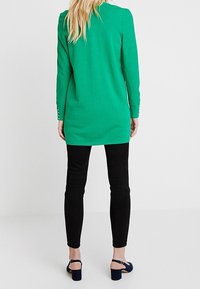 Zelená tunika s dlouhým rukávem a kulatým výstřihem, doplněná o knoflíkové detaily na rukávech, v kombinaci s černými slim kalhotami a modrými vysokými podpatky.