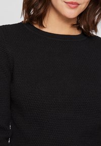 Μαύρο πλεκτό πουλόβερ με υφή, στρογγυλή λαιμόκοψη και ρέσες στους καρπούς. Το ύφασμα έχει μαλακό, ελαφρώς ανυψωμένο πλέγμα.