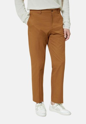 Mand iført brune skræddersyede bukser med sidelommer og hvide lavtop-sneakers, stående med den ene hånd i lommen.