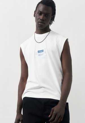 Bărbat de culoare cu dreadlocks scurte, purtând un tricou alb fără mâneci HUGO și un colier argintiu cu lanț, pe un fundal simplu, deschis la culoare.