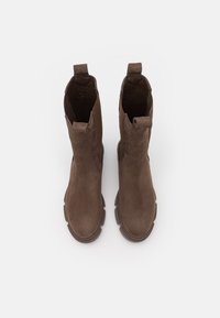Bottes de cheville en daim marron avec une surface lisse, des panneaux élastiques sur les côtés et des languettes de tirage. La semelle présente des bords arrondis et une finition texturée.